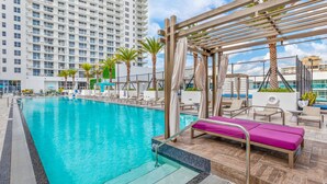 A swimming pool with lounge chairs and a pergola, surrounded by palm trees and a high-rise building. - OCEAN VIEW 2BD-2BA CONDO 2207 (Hollywood)