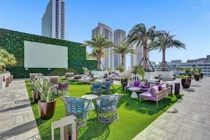Um lounge no terraço com assentos externos, palmeiras e vista para edifícios altos.