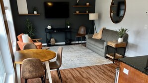 A modern living room with a wooden floor, a grey sofa, a dining table with chairs, a TV mounted on the wall, and a small side table with a lamp. - Pristine Condo, Close to Downtown, Beaches, GT Commons, and Restaurants (Traverse City)
