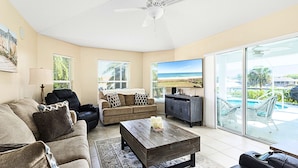 A spacious living room with a large TV, a ceiling fan, and a view of a pool and beach through large windows. - Dock Holliday by Ryson Vacation Rentals (Palm Coast)