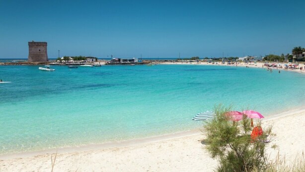 A beach with clear turquoise water, a sandy shore, and a tower in the background.