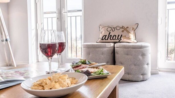 A dining area with a wooden table, two glasses of red wine, a bowl of chips, a plate with a salad, and a grey ottoman with a decorative pillow. - Rest Ashored, Aldeburgh (Aldeburgh)