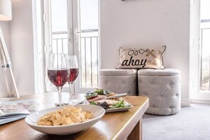 A dining area with a wooden table, two glasses of red wine, a bowl of chips, a plate with a salad, and a grey ottoman with a decorative pillow. - Rest Ashored, Aldeburgh (Aldeburgh)