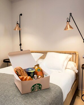 Una habitación de hotel con una cama, una mesita de noche, una lámpara y una caja de productos de Starbucks.