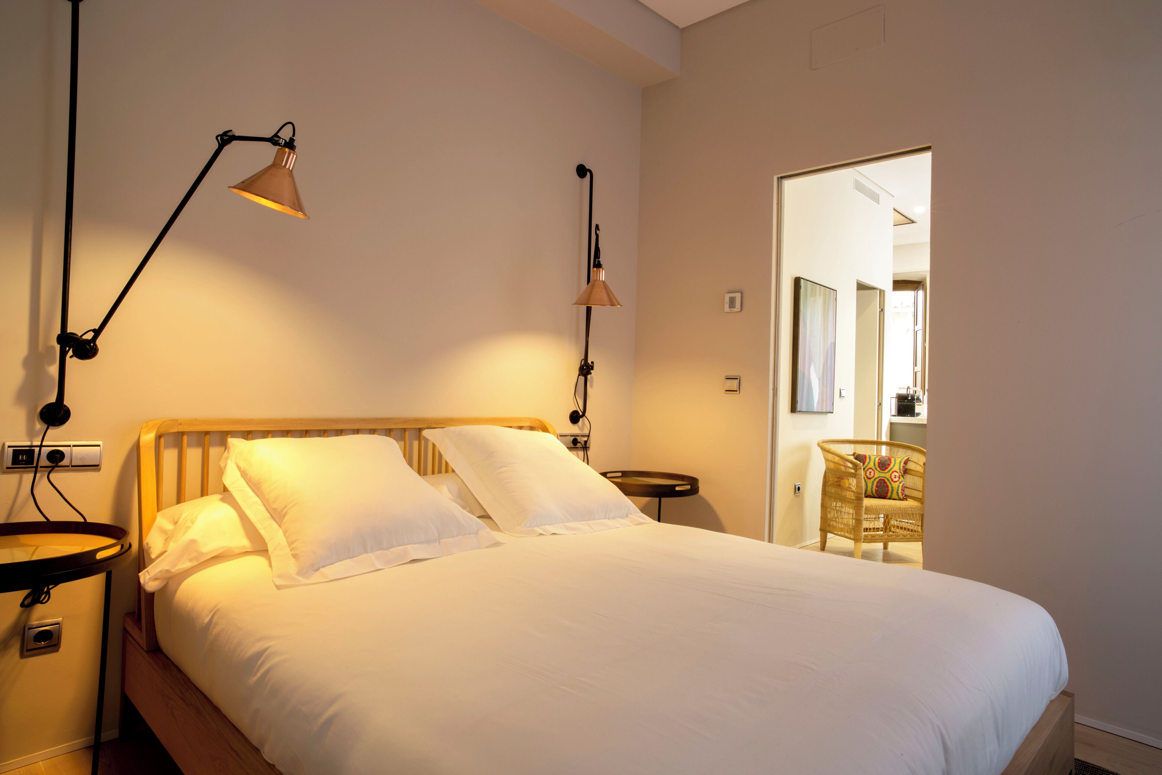 Una cama bien hecha con sábanas blancas, dos lámparas de noche, un cabecero de madera y una silla de mimbre al fondo.