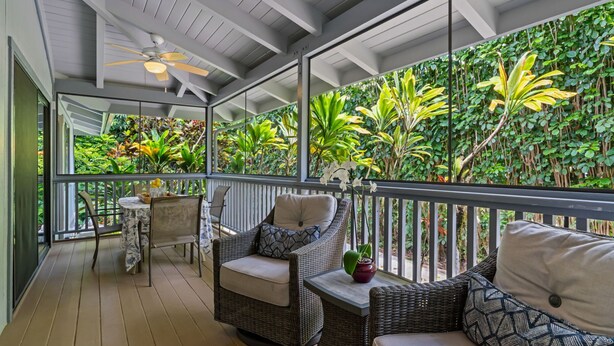 A covered outdoor area with wicker furniture, a ceiling fan, and a view of lush greenery through large windows.