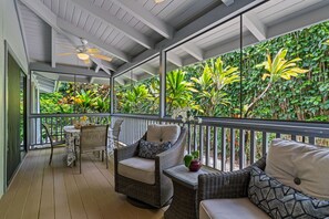 Un espace extérieur abrité, aménagé avec des meubles en osier, un ventilateur de plafond et offrant une vue sur une végétation luxuriante grâce à de grandes fenêtres.