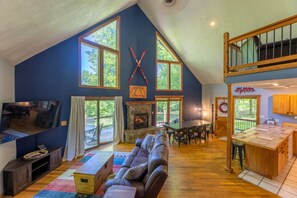 A spacious living area with a stone fireplace, large windows, a high ceiling, and a cozy seating arrangement.