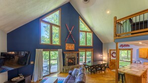 A spacious living area with a stone fireplace, large windows, and a cozy seating arrangement. - Base Camp - Cozy Cabin, Close to Snowshoe! (Pocahontas County)