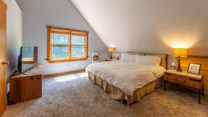 A bedroom with a sloped ceiling, a bed, a nightstand, a lamp, a TV, and a window. - Base Camp - Cozy Cabin, Close to Snowshoe! (Pocahontas County)