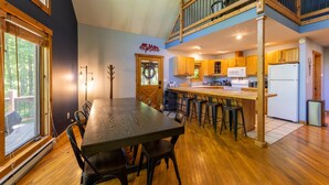 A spacious kitchen with wooden cabinets, a central island with stools, and a view of the outdoors through large windows. - Base Camp - Cozy Cabin, Close to Snowshoe! (Pocahontas County)