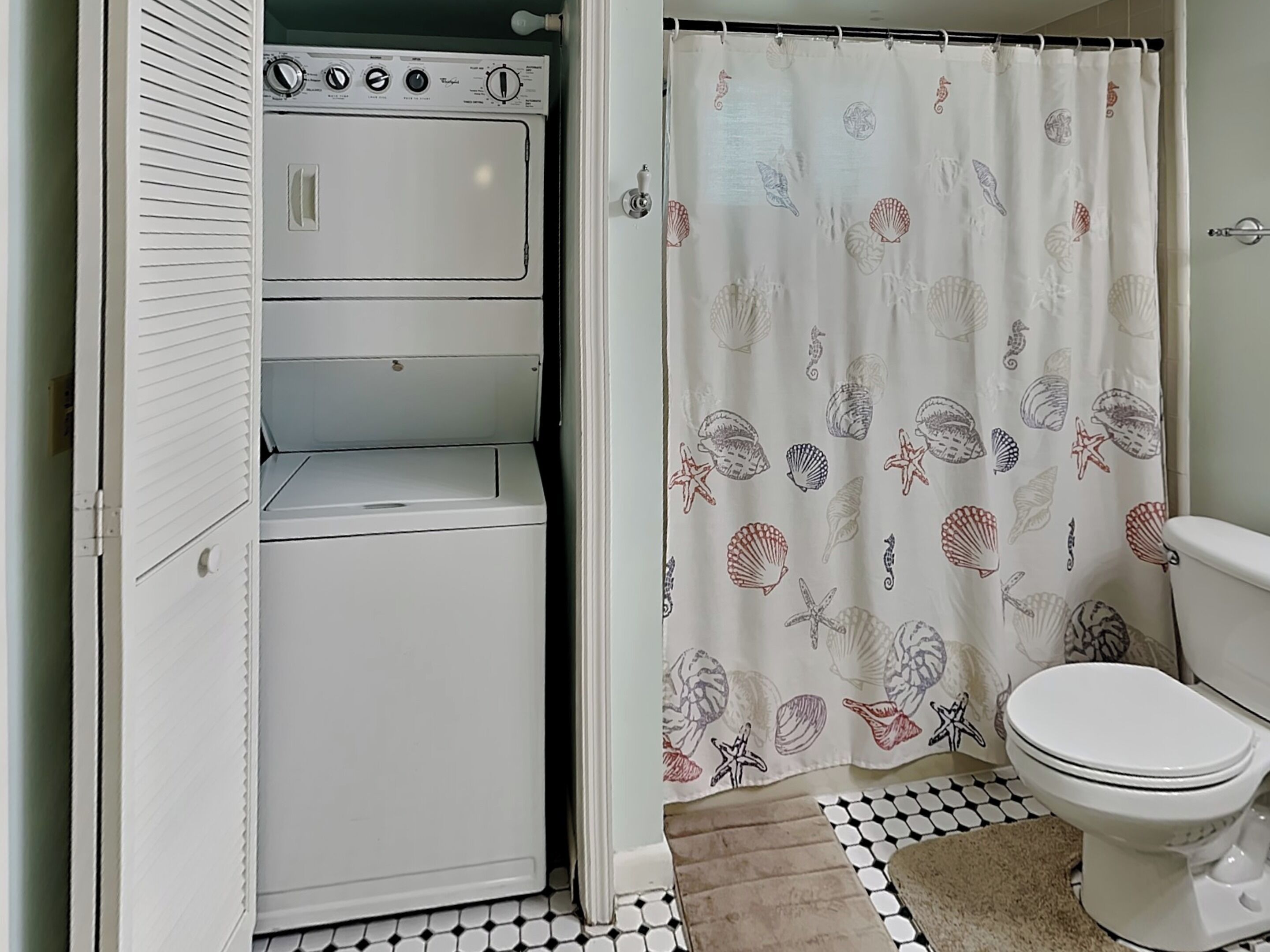 A compact laundry room with a washing machine and dryer, a shower curtain with a sea shell pattern, and a toilet.