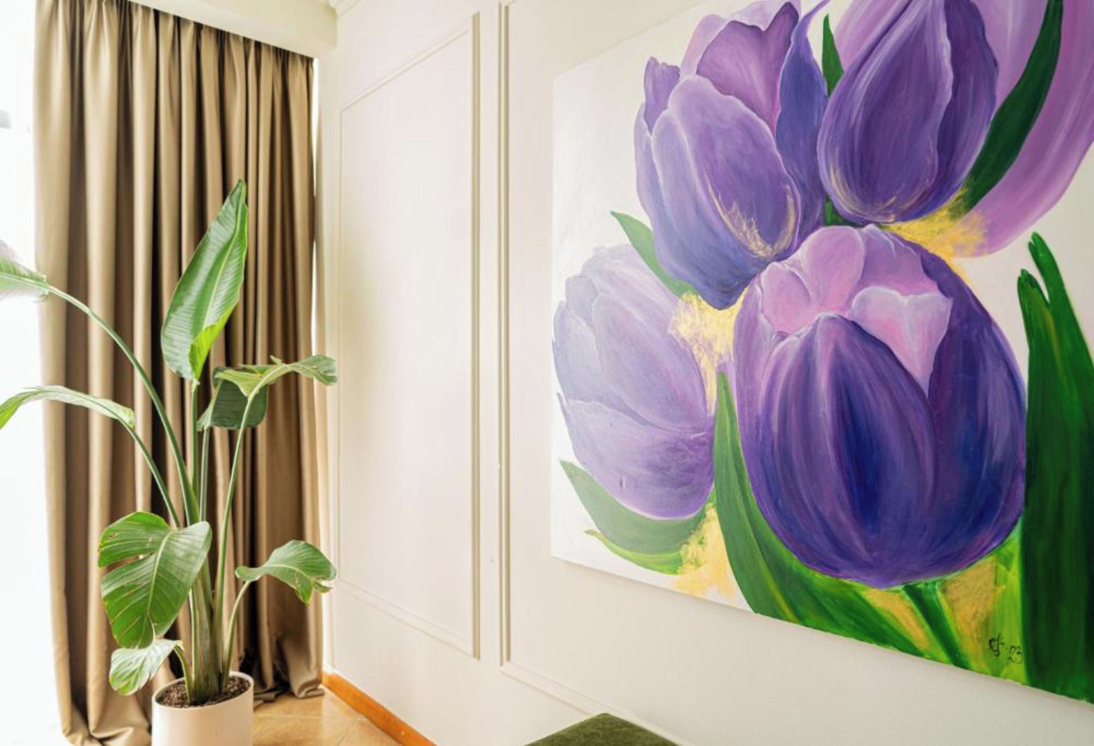 A room with a large potted plant, a painting of purple flowers on the wall, and a window with curtains.