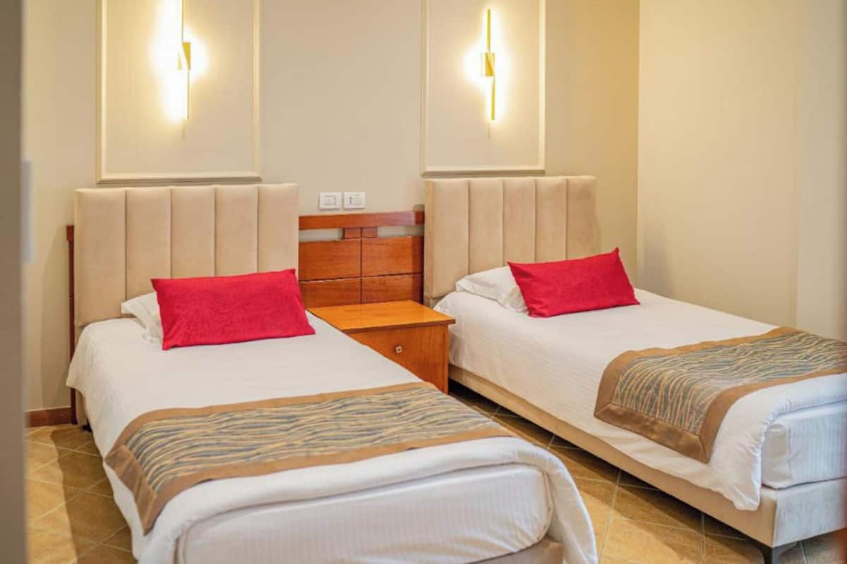 A hotel room with two beds, each with a red pillow and a striped blanket. There are wall-mounted light fixtures above the beds.
