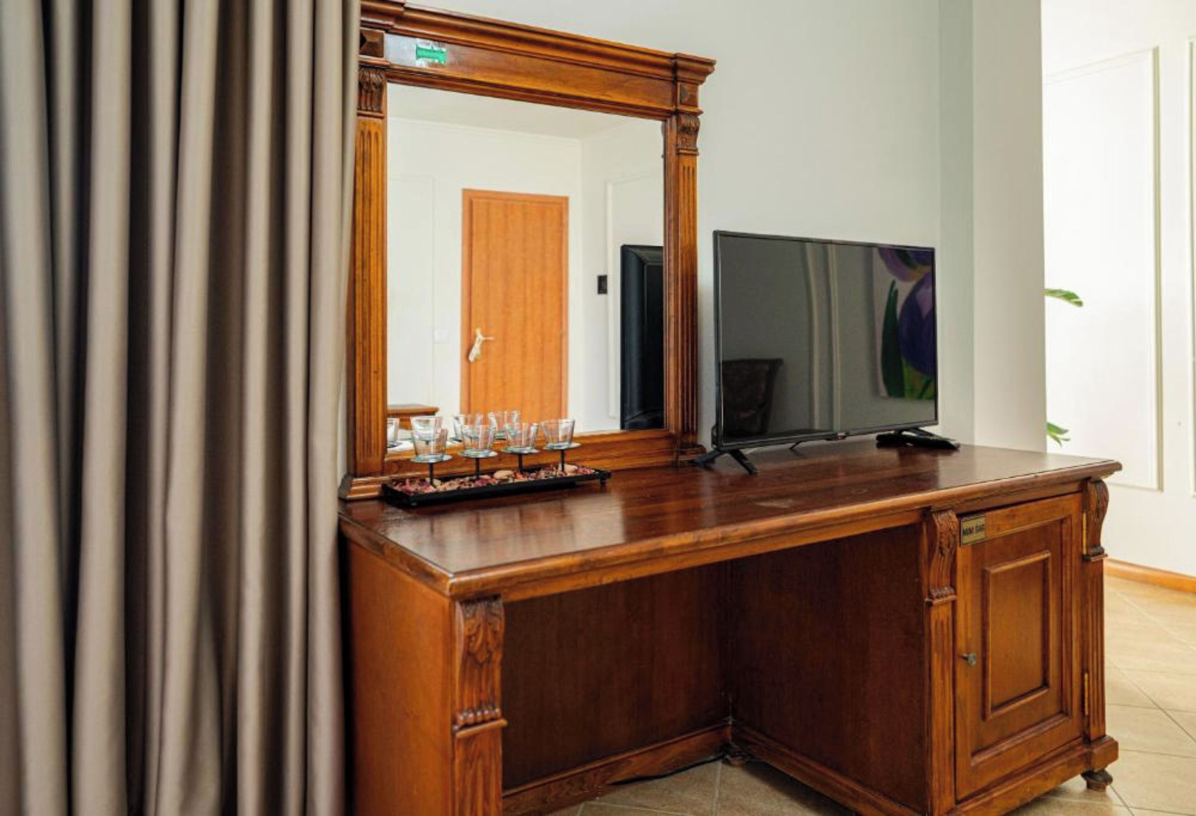 A wooden desk with a mirror, a flat-screen TV, and a tray with glasses and a bottle.