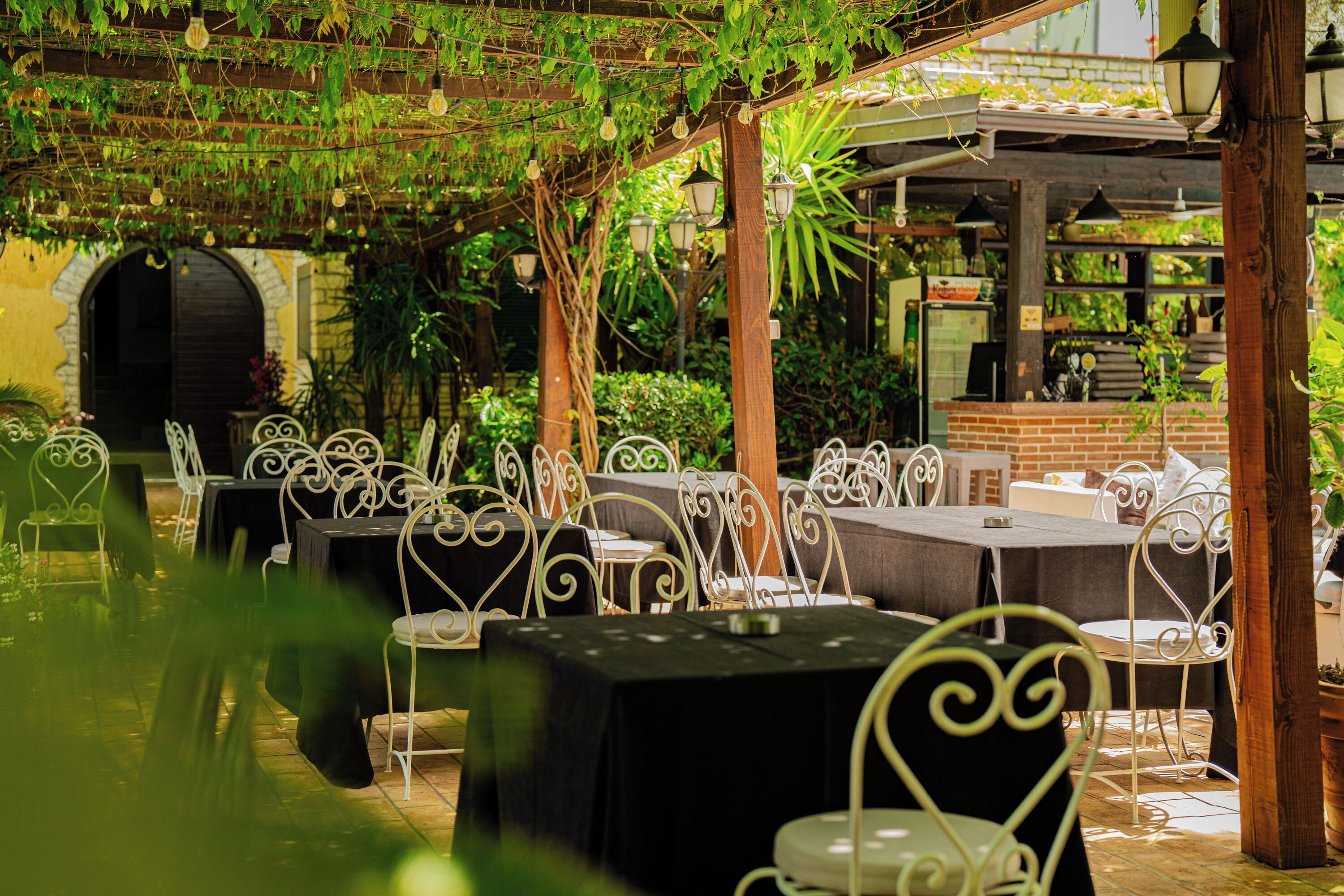 An outdoor dining area with tables and chairs, surrounded by greenery and a brick wall.