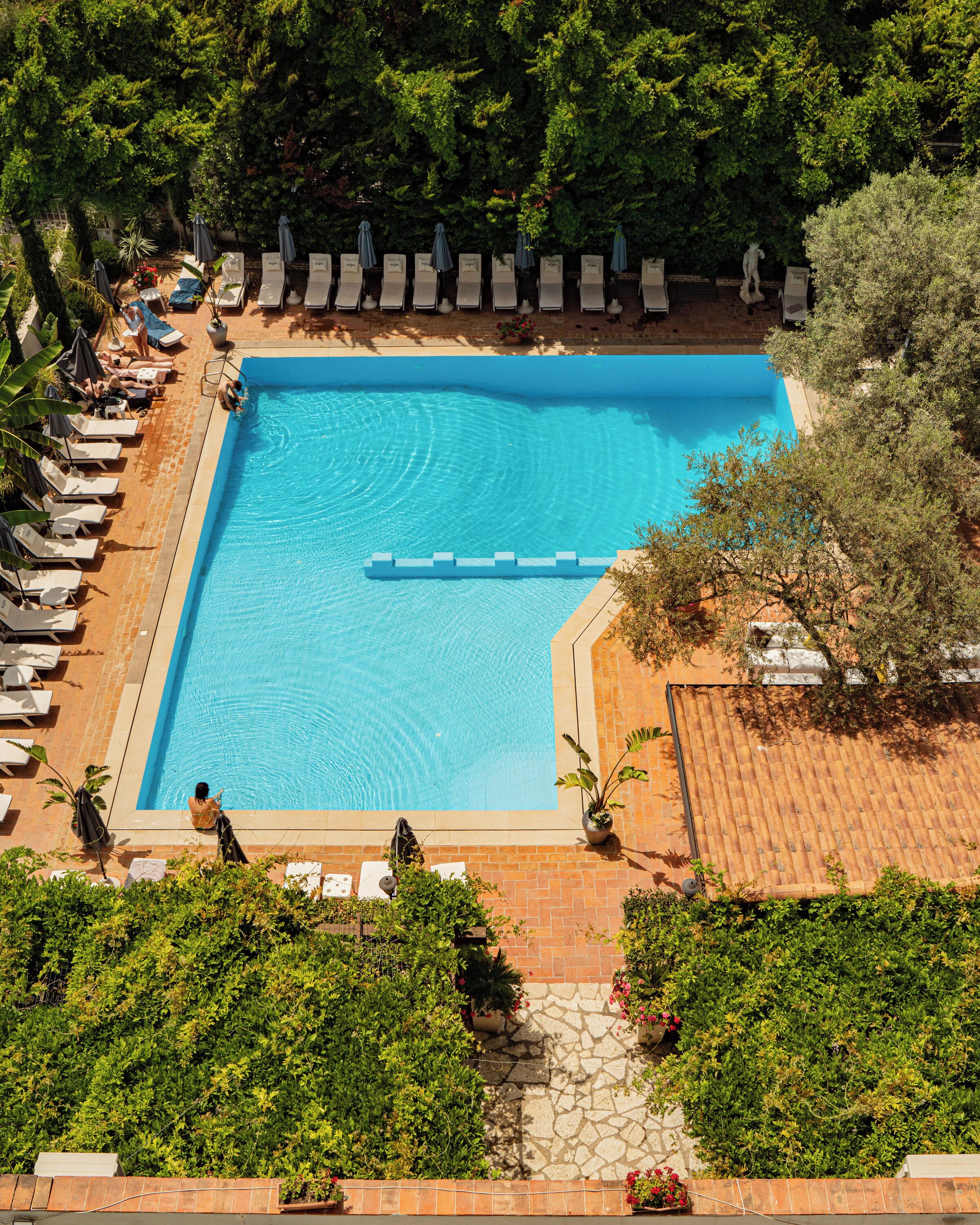 A rectangular swimming pool surrounded by lounge chairs and umbrellas, with a stone wall and greenery in the background.