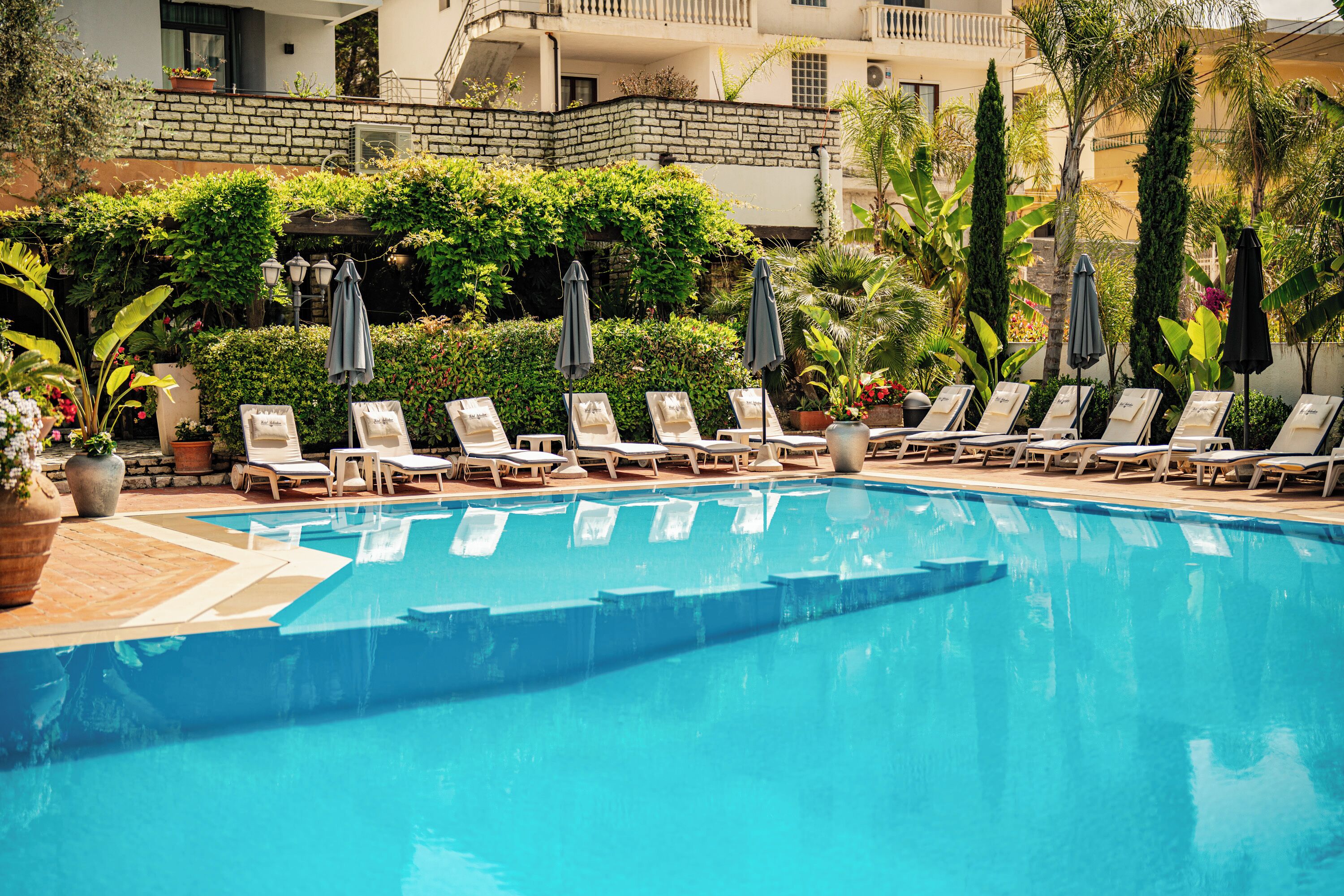 Piscina con sillas de descanso y sombrillas, rodeada de vegetación y un edificio al fondo.