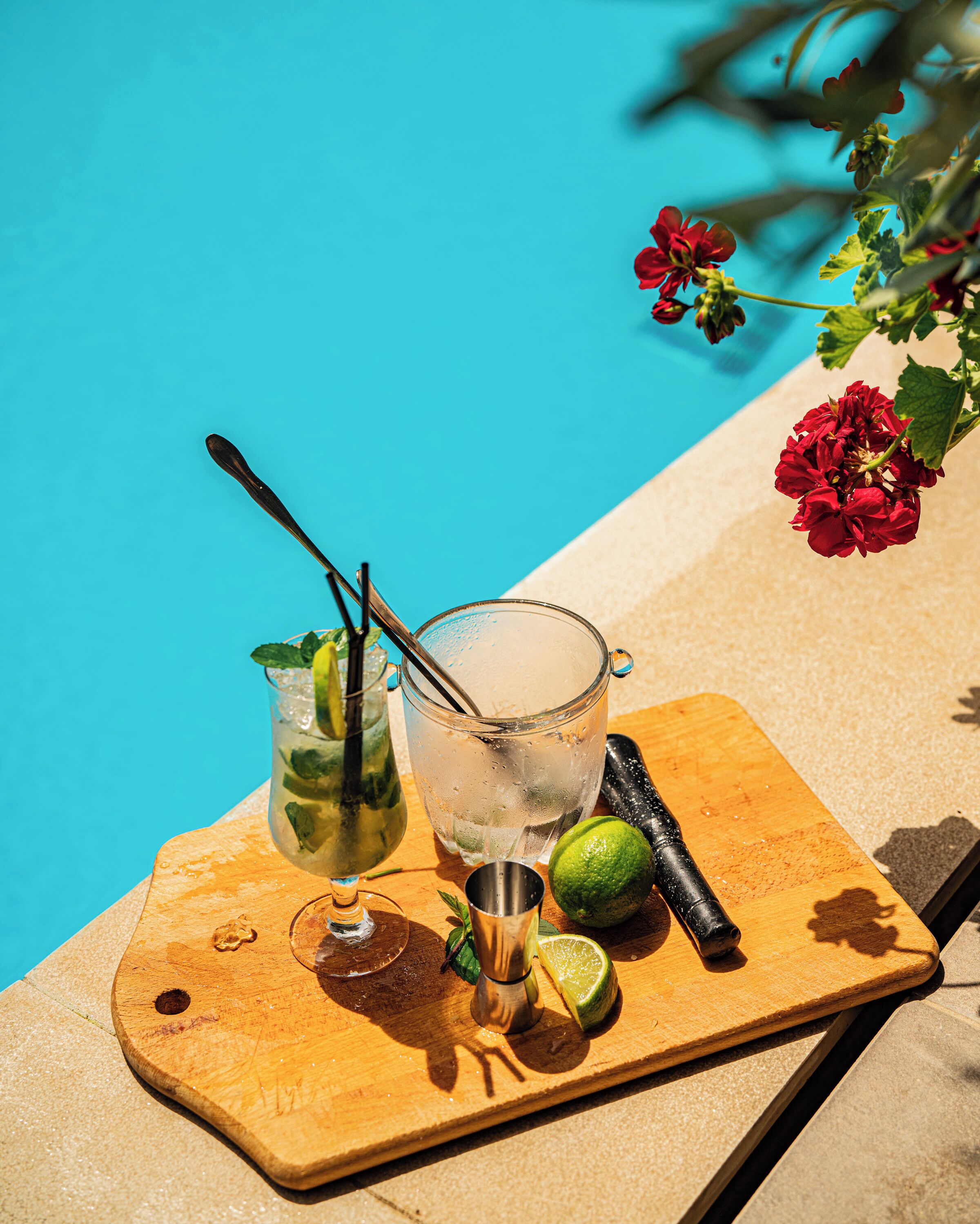 Una taula de fusta amb un got de beguda gelada, un agitador de còctels i un llimona a plena sol al costat d'una piscina.