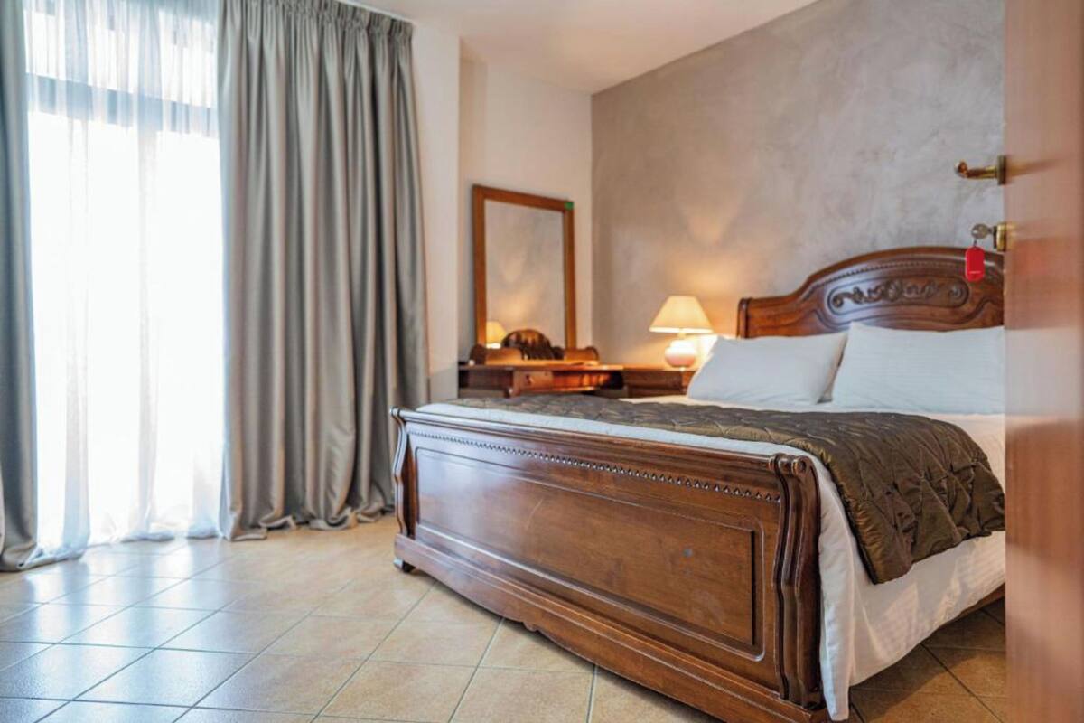 A hotel room with a large bed, a nightstand, a lamp, a mirror, and a window with curtains.