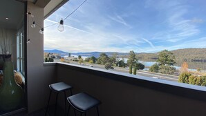 A balcony with a view of a lake, mountains, and a road. There are two black stools and a hanging light fixture. - Jasper 4 in the Town Centre with Lake views (Jindabyne)