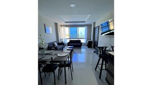 A modern living room with a sofa, dining table, and chairs, a kitchen area with a TV, and large windows offering a view. - Oceanfront Getaway • Nature • Comfort • Gated Community / Frente al Mar (General Villamil Playas)