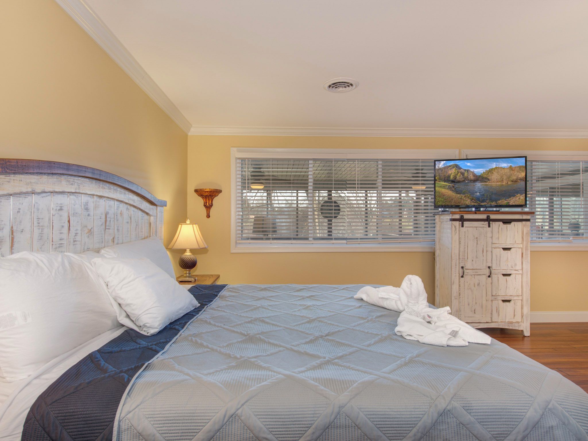 A bedroom with a large bed, a television on a stand, a window with blinds, and a wooden cabinet.