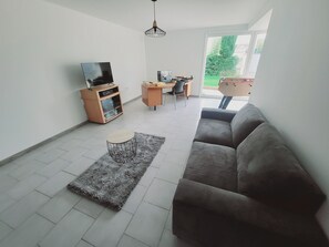 Un salon moderne avec un canapé gris, une petite table basse ronde, un téléviseur posé sur un meuble en bois, un bureau avec une chaise et une grande fenêtre donnant sur un paysage verdoyant.