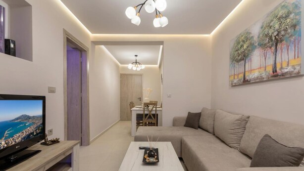 A modern living room with a flat-screen TV, a white coffee table, a beige sofa, and a dining area in the background. - Evia s Apartment - New Apartment In Town - Evia s Apartment - New Apartment In Town (Zakynthos)