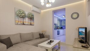 A modern living room with a sofa, a coffee table, and a TV. There is a painting on the wall and a kitchen visible through an open doorway. - Evia s Apartment - New Apartment In Town - Evia s Apartment - New Apartment In Town (Zakynthos)