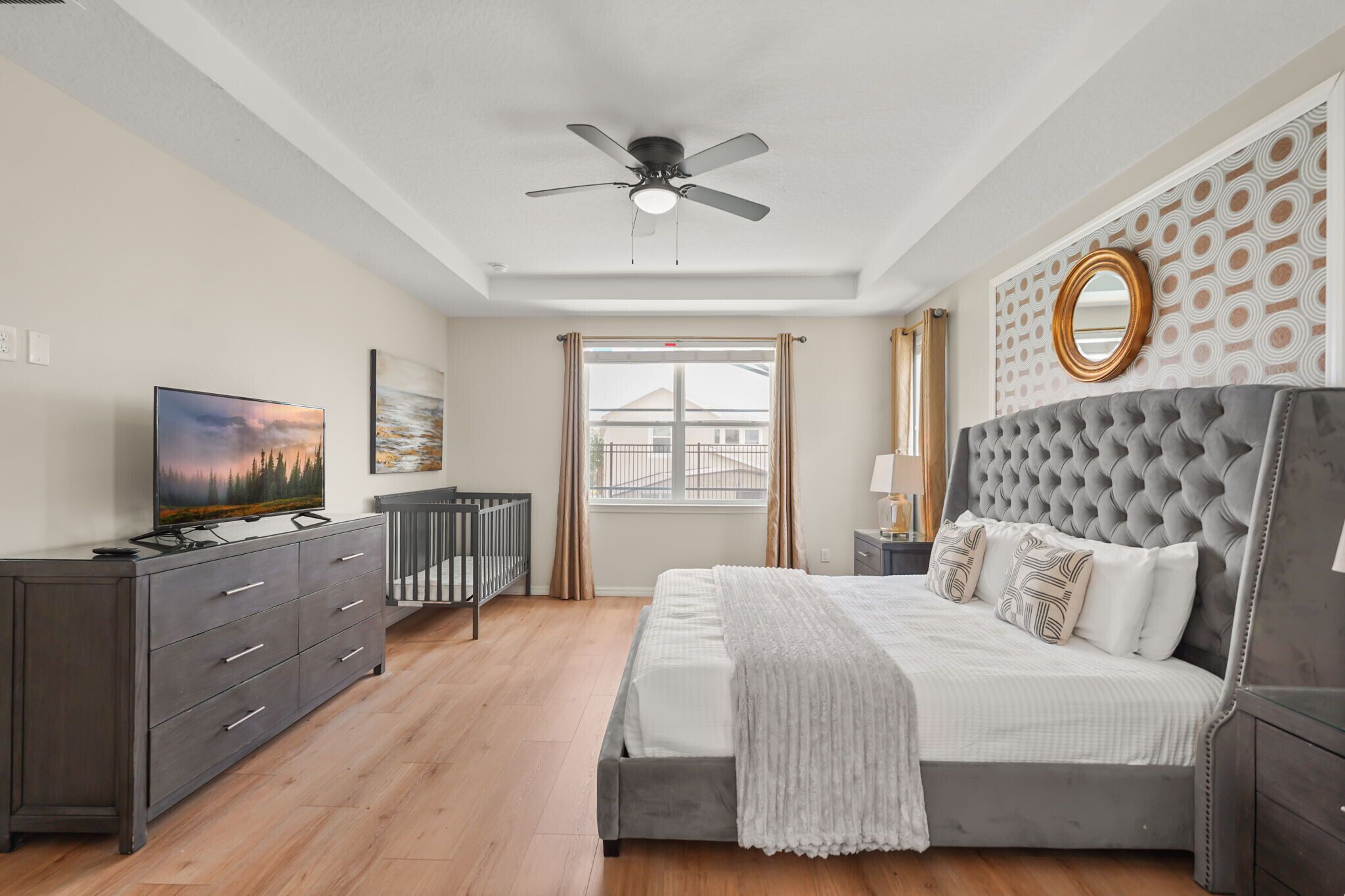 Une chambre à coucher comprenant un lit, un ventilateur de plafond, une télévision, une commode, un berceau et une fenêtre avec des rideaux.