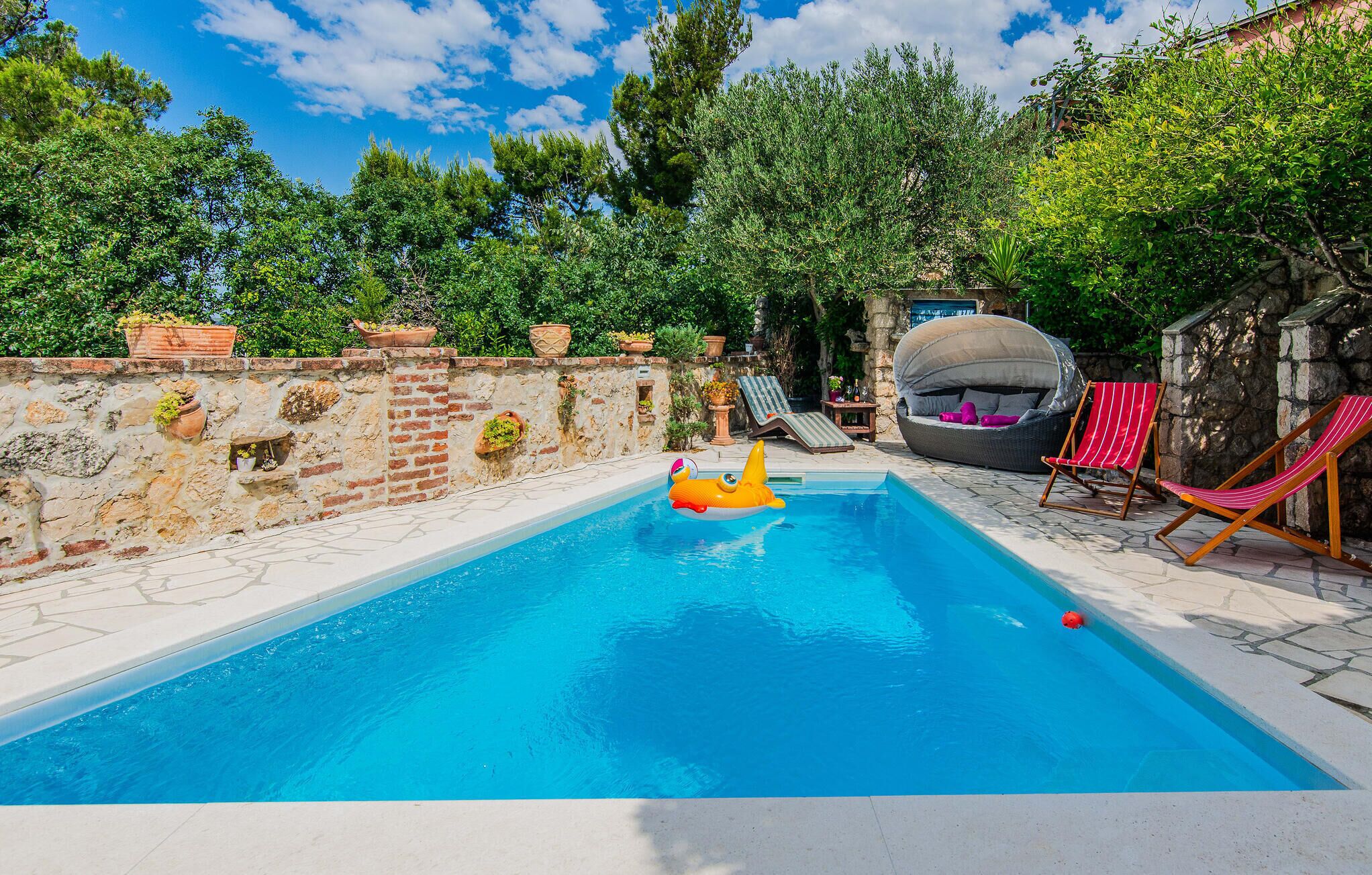 A swimming pool with a stone wall, a lounge chair, and a covered seating area.