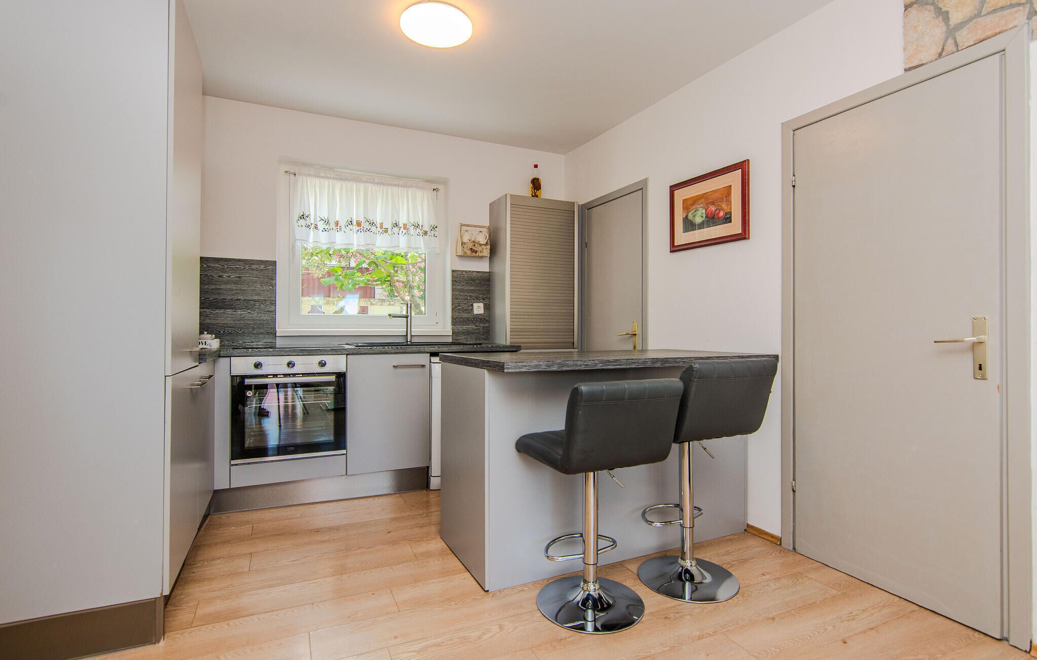 A compact kitchen with a built-in oven, a window with curtains, and a small bar area with two stools.