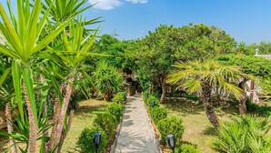 Un sentier de jardin bordé de diverses plantes et arbres tropicaux, menant à une construction en pierre.