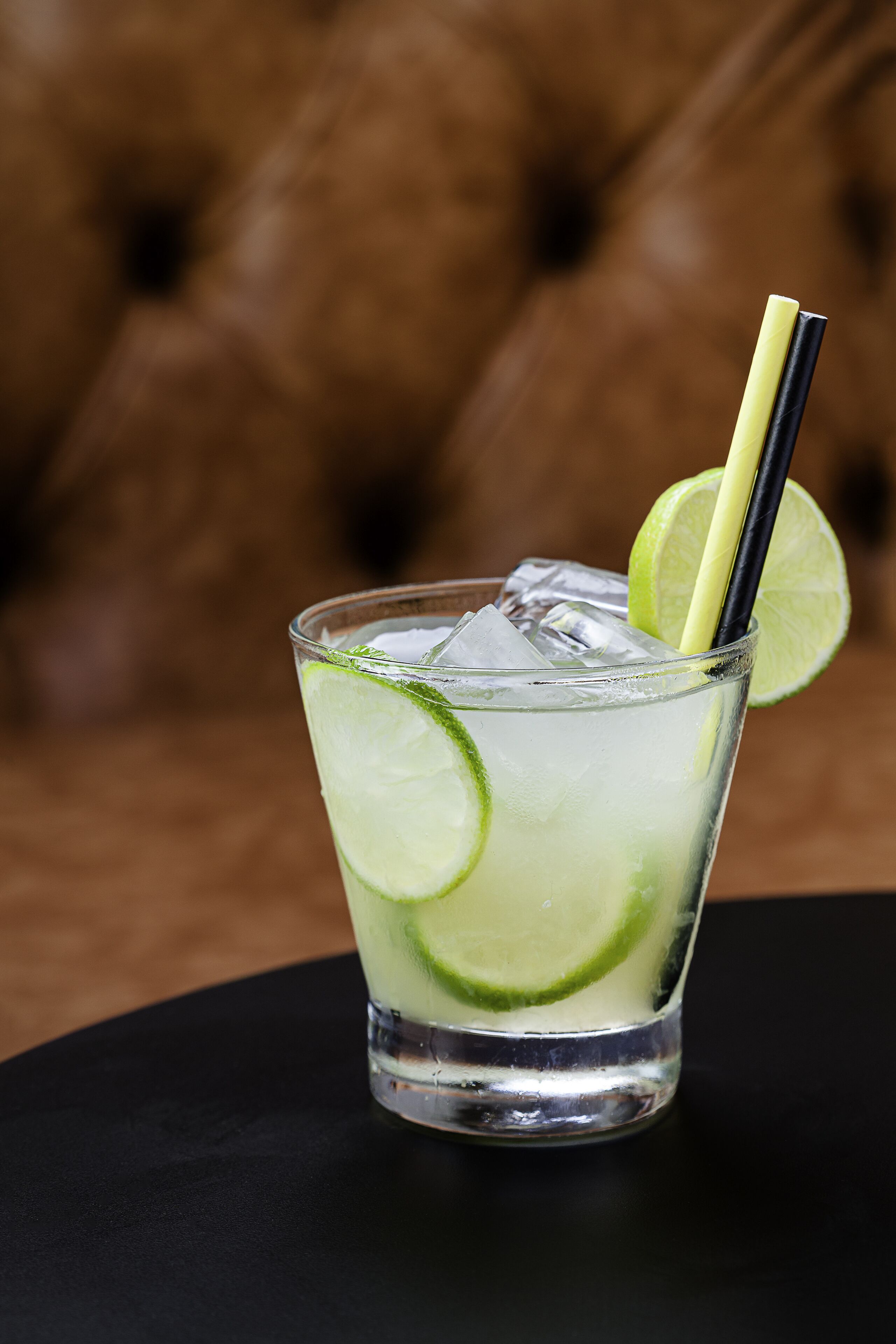 A glass of a clear beverage with lime slices and a straw, placed on a black surface with a brown leather couch in the background.