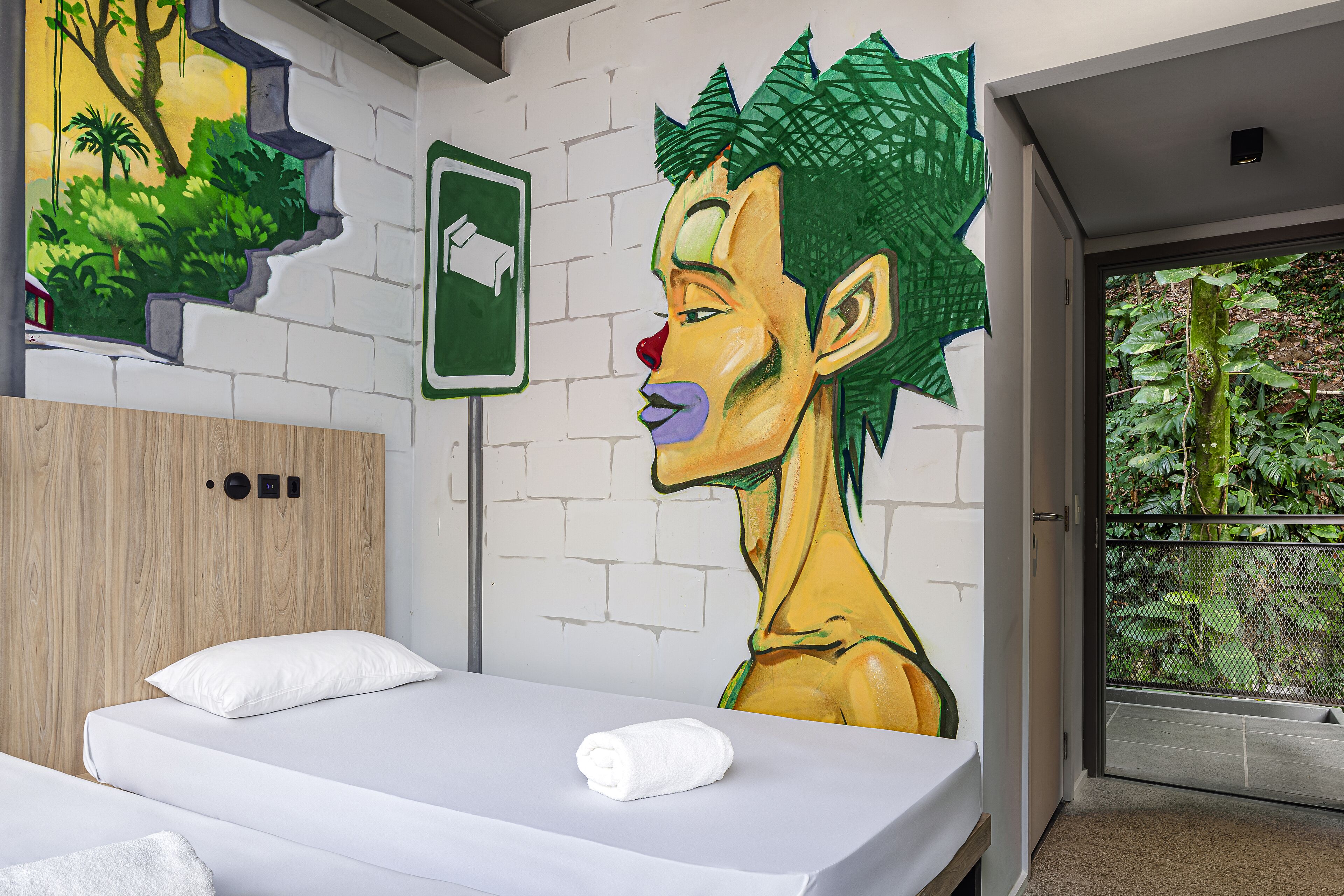 A hotel room with a bed, a white pillow, a green wall mural, and a window with a view of greenery.