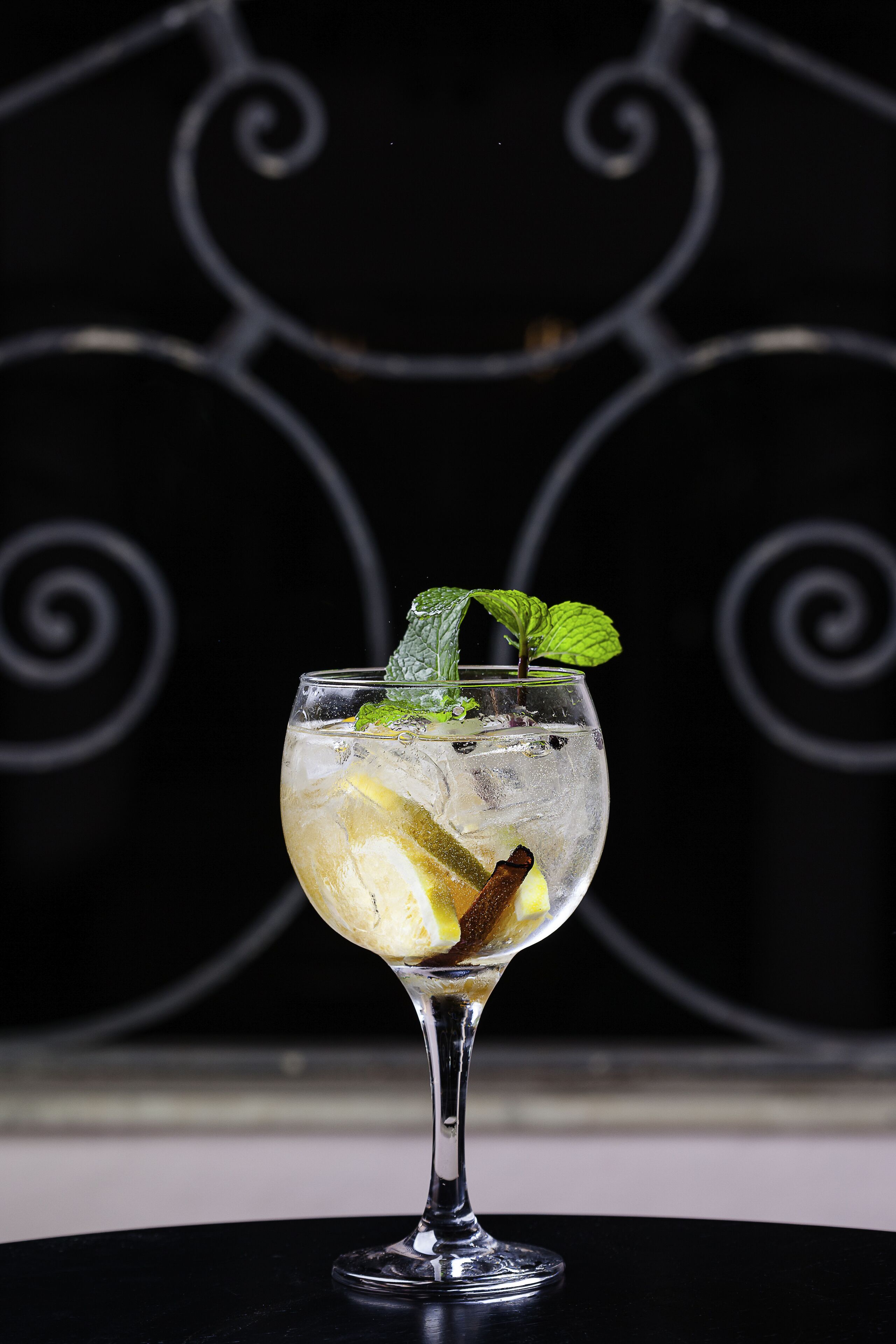 A cocktail glass with ice, lemon slices, mint leaves, and a cinnamon stick on a black table with a decorative metal background.