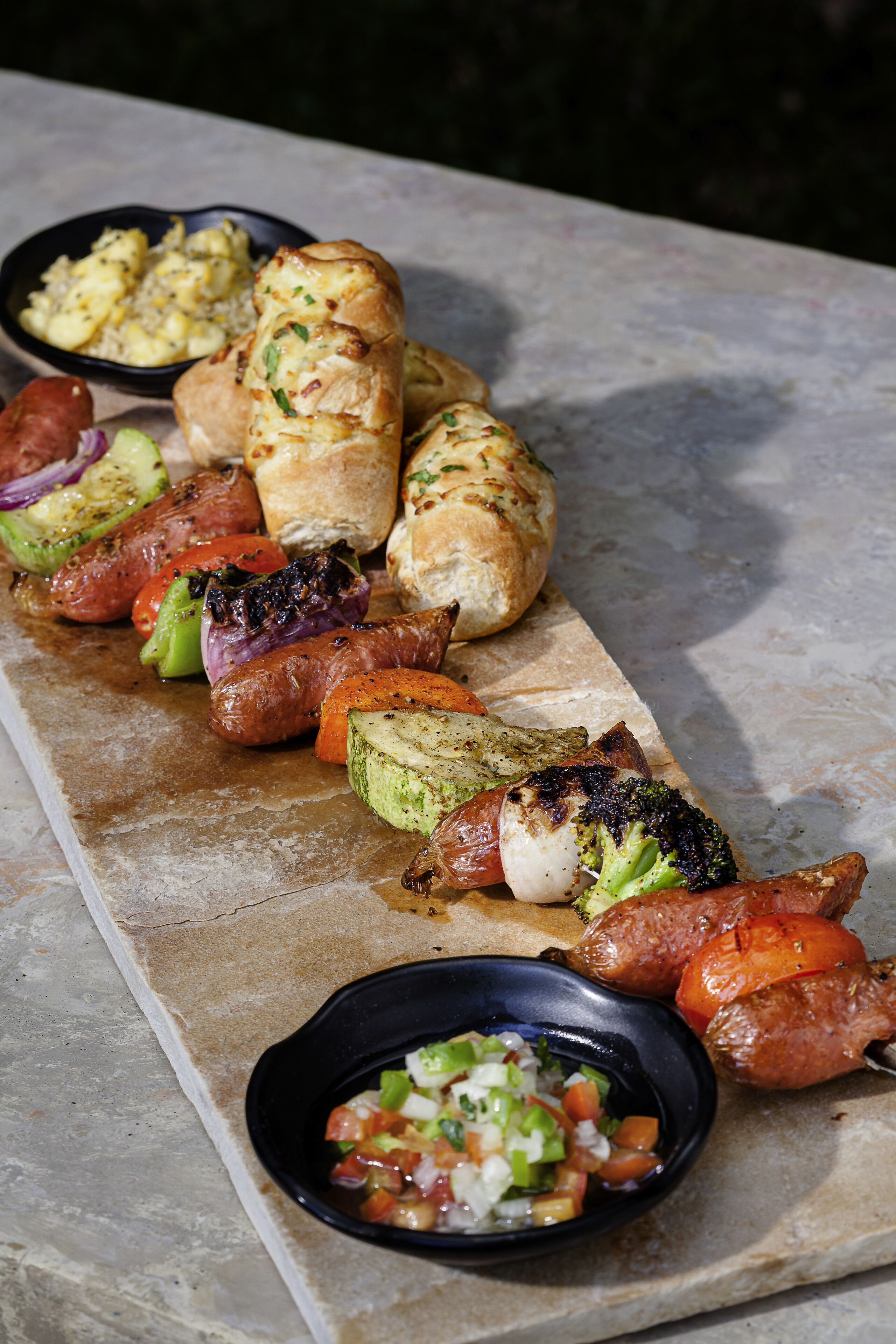 Una mesa de piedra con salchichas a la parrilla, verduras y pan.
