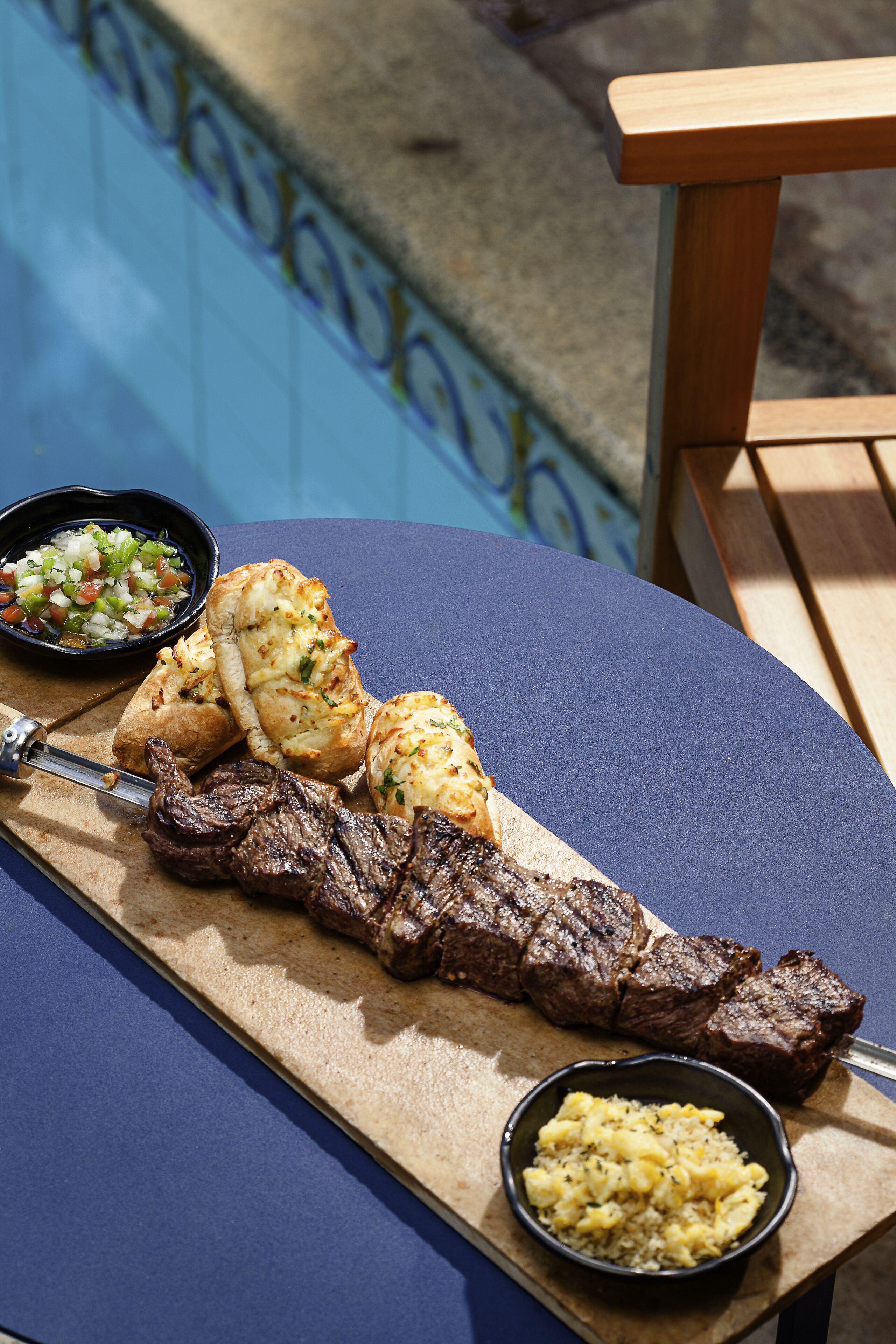 A poolside table with a skewer of grilled meat, garlic bread, and two small bowls of side dishes.