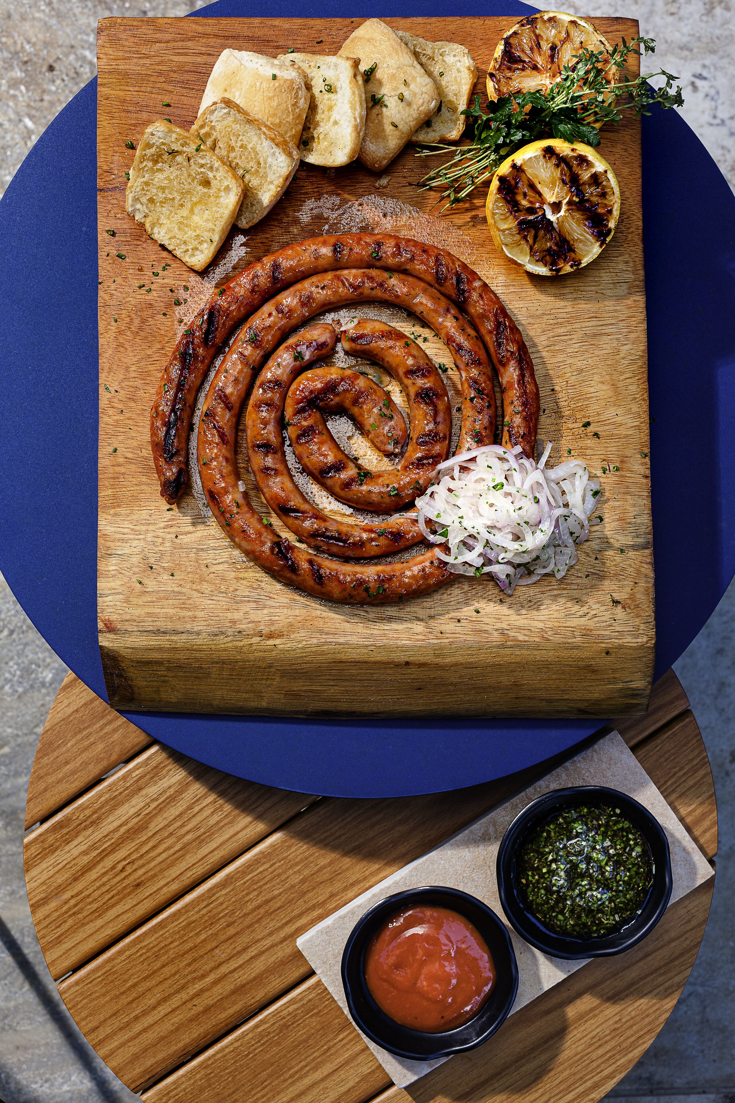 A wooden cutting board with grilled sausages, sliced bread, grilled lemon, and onions.