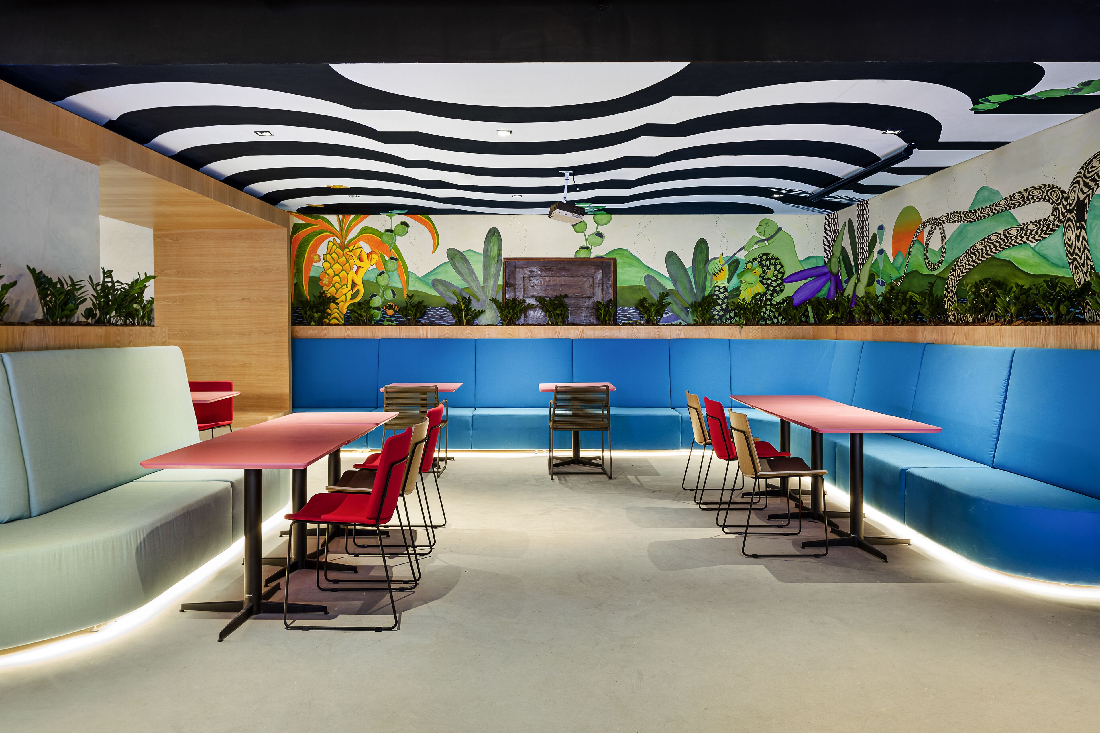 Een modern restaurant met blauwe zitplaatsen, rode tafels en een muurschildering van tropische planten.