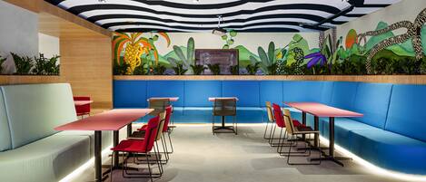 Een modern restaurant met blauwe zitplaatsen, rode tafels en een muurschildering van tropische planten.