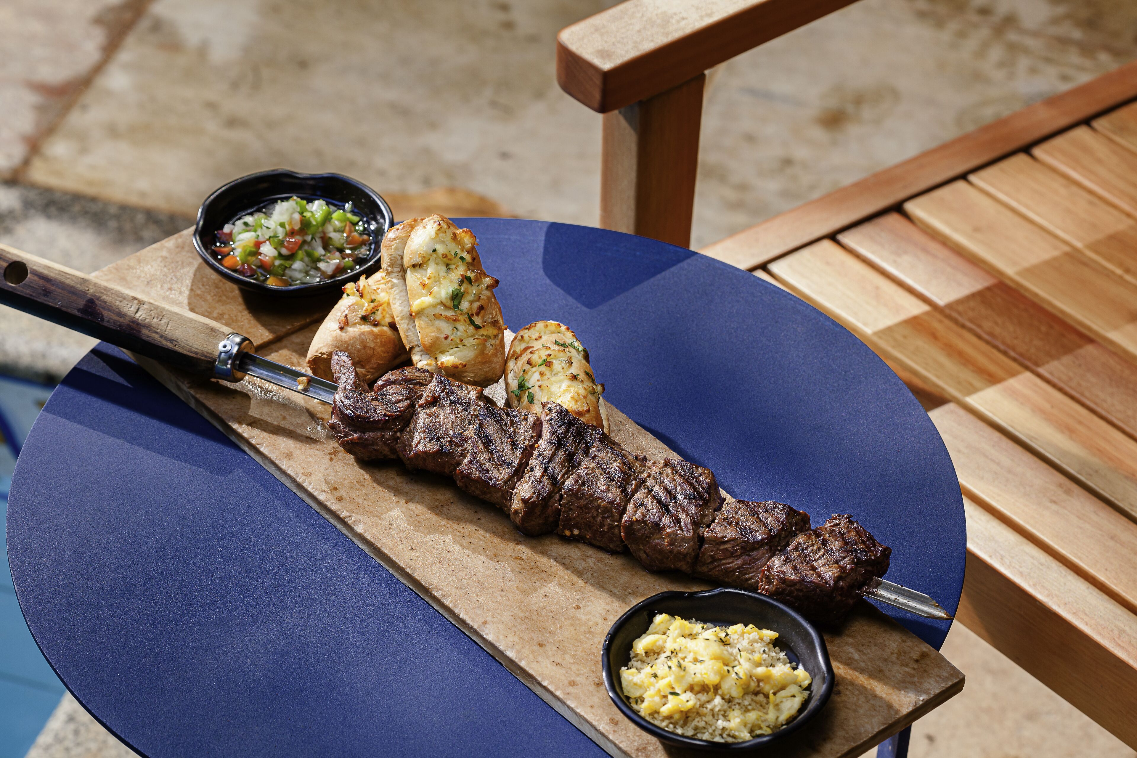 A wooden table with a blue surface, a skewer of grilled meat, and two small bowls of side dishes.