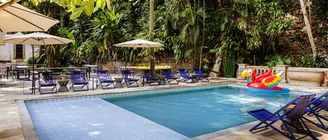A swimming pool with tiled flooring, surrounded by blue lounge chairs and umbrellas, set against a backdrop of dense foliage.