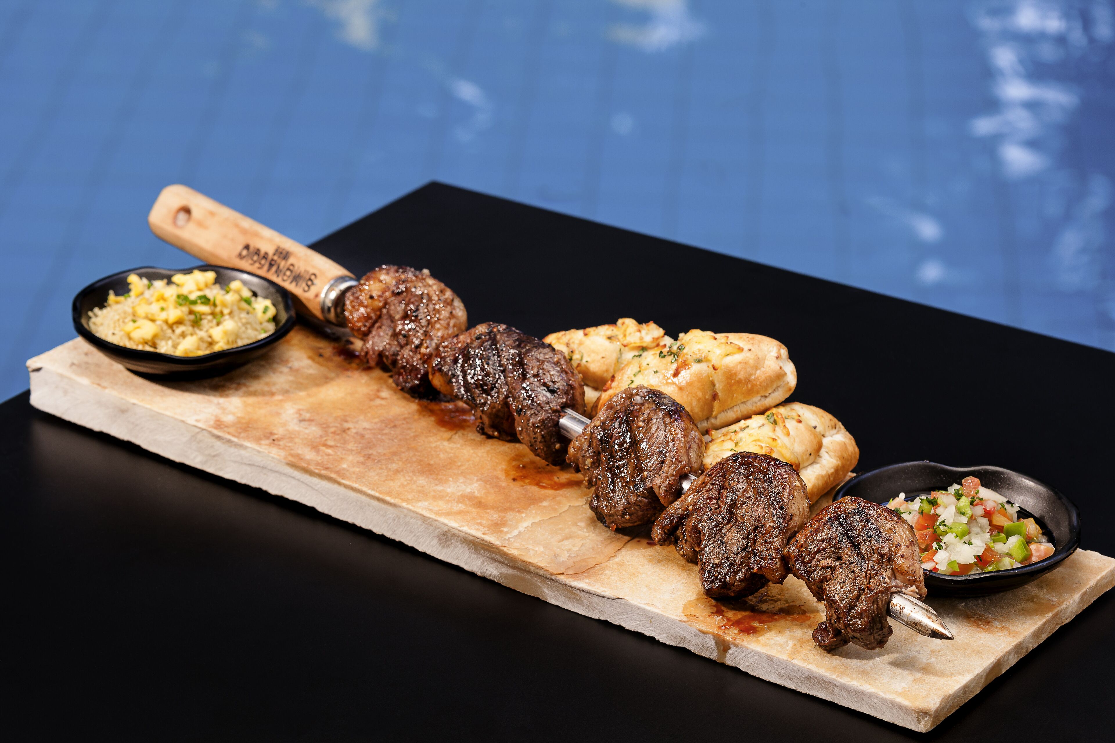 Una tabla de madera con carne ensartada, panes planos y dos pequeños recipientes de acompañamientos.