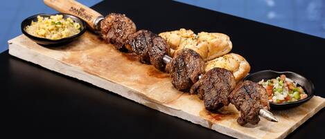 Una tabla de madera con carne ensartada, panes planos y dos pequeños recipientes de acompañamientos.