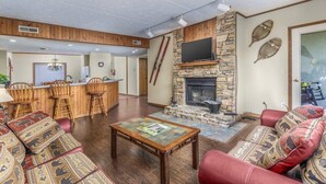 A living room with a stone fireplace, wooden furniture, and a kitchen area in the background.