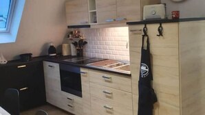 Eine kompakte Küche mit hellen Holzschränken, einer schwarzen Arbeitsplatte und einem eingebauten Backofen. An der Wand hängt eine Uhr und ein Fenster sorgt für natürliches Licht.