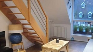 Ein modernes Wohnzimmer mit einer Holztreppe, einem großen Fenster mit Blick auf Häuser, einem kleinen Tisch und einem gelben Sessel.