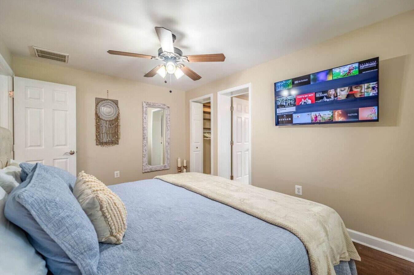 Una camera da letto con un letto, una ventola a soffitto, una TV a muro che mostra diversi canali e una porta che dà su un'altra stanza.